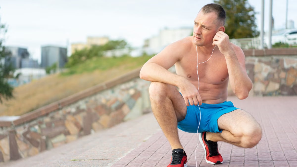Shirtless muscular man in blue shorts and red shoes listening to music outdoors.